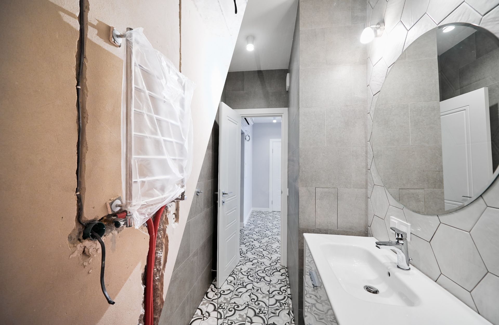 Narrow bathroom under renovation with exposed plumbing, gray walls, hex tile floor, and a white sink mirror.
