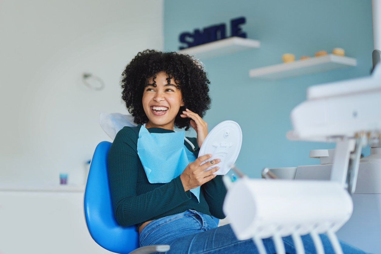 A woman is sitting in a dental chair looking at her teeth in a mirror.