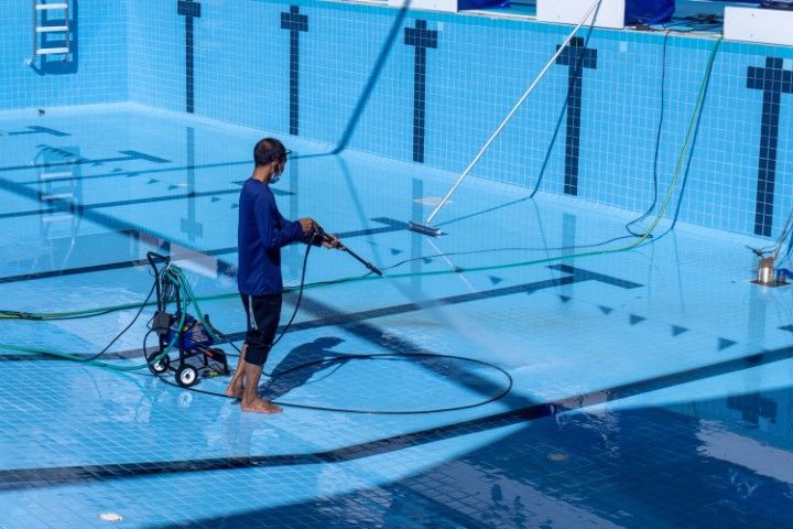 Person cleaning a blue tiled swimming pool with a pressure washer.