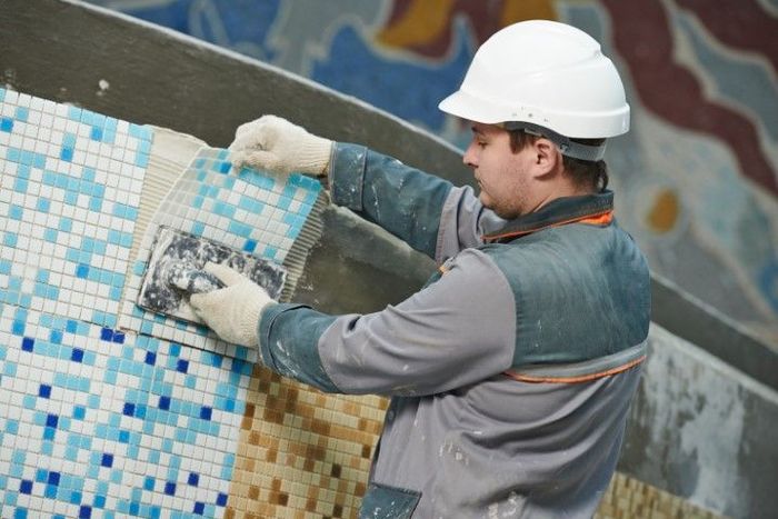 Construction worker in white hard hat applying grout to mosaic tiles on a curved wall.