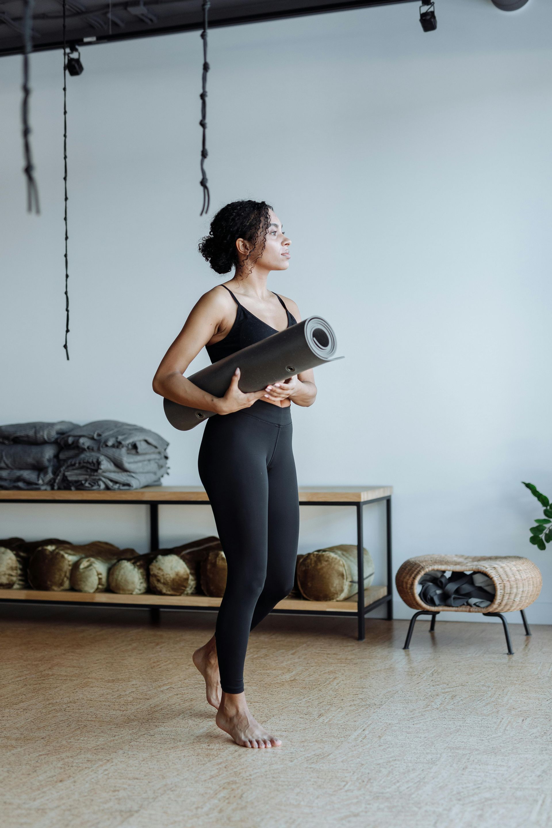 Mujer vestida con ropa deportiva negra llevando una esterilla de yoga en un estudio.