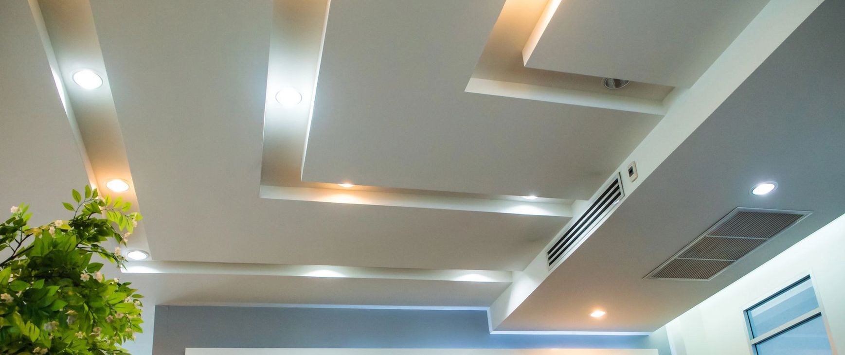 Soffitto interno moderno con faretti a incasso, finiture beige, bocchette di ventilazione e una pianta in vaso a sinistra.