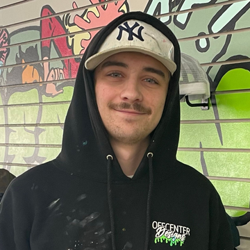 Man in a black hoodie and Yankees cap smiles, posing in front of a colorful graffiti wall.