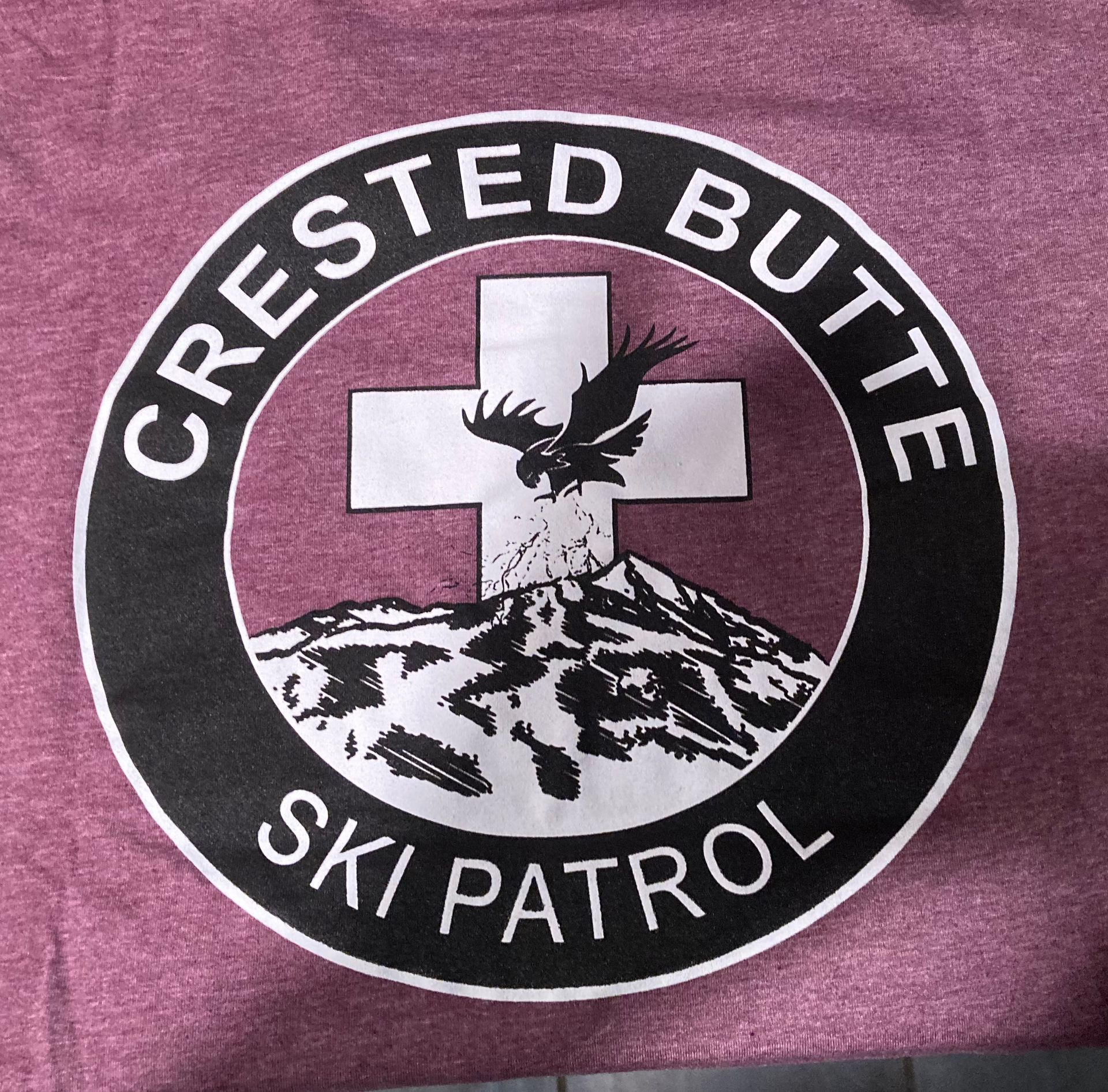 Crested Butte Ski Patrol logo on maroon fabric; a black circle with white lettering, cross, eagle and mountain.