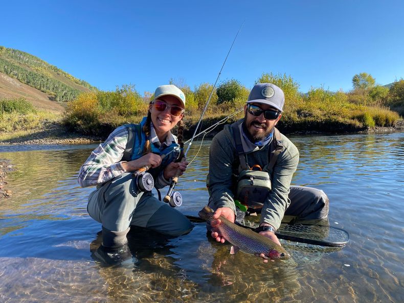 Two people, one holding a fishing rod, the other holding a fish in a river, both smiling. Sunny, outdoor setting.
