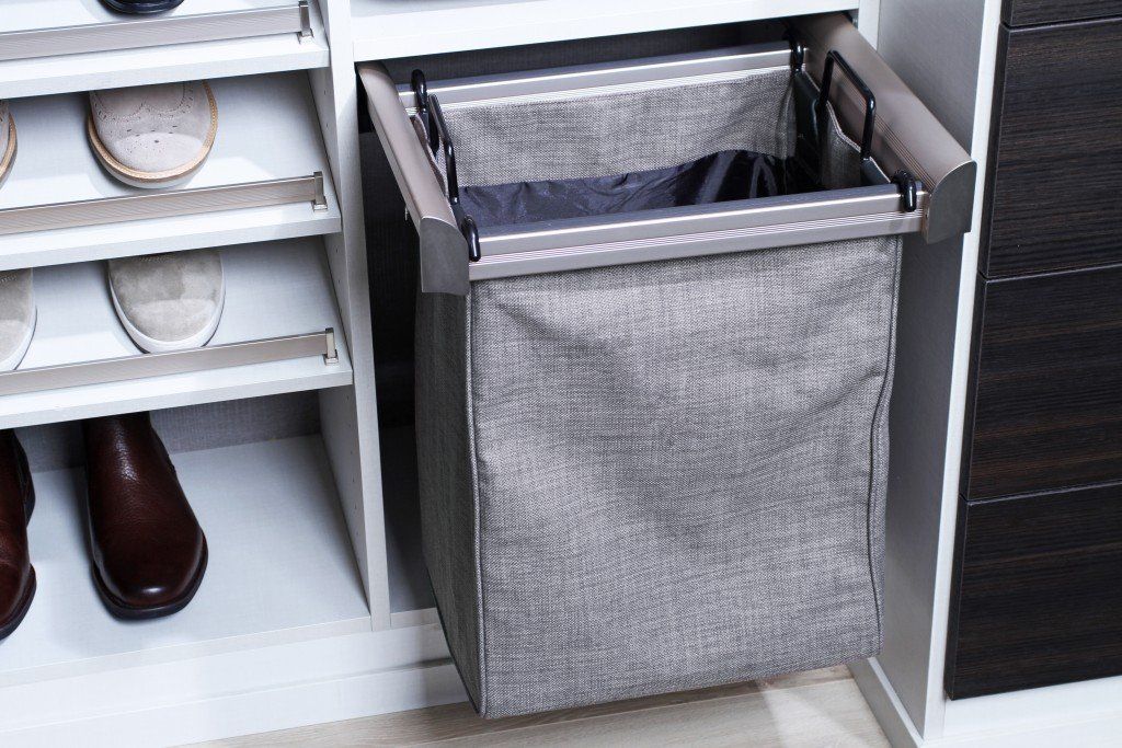 Gray fabric pull-out hamper in a closet beside white shoe shelves and dark shoes