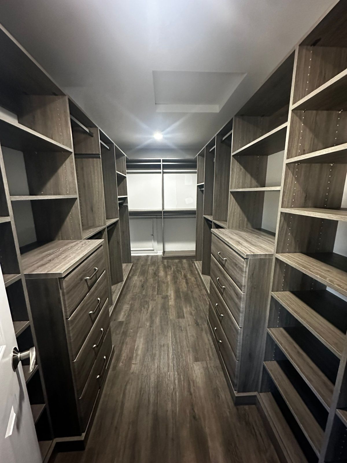 Walk-in closet with dark wood shelves, drawers, and flooring. Empty, with a center aisle and a light in the ceiling.