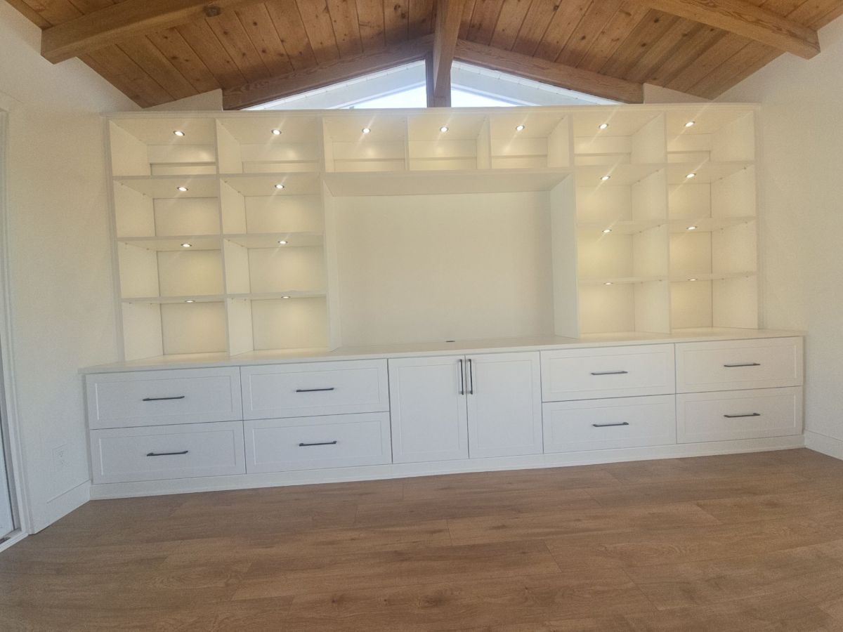 White built-in wall unit with drawers and shelves, lights on, against a white wall and wood ceiling.