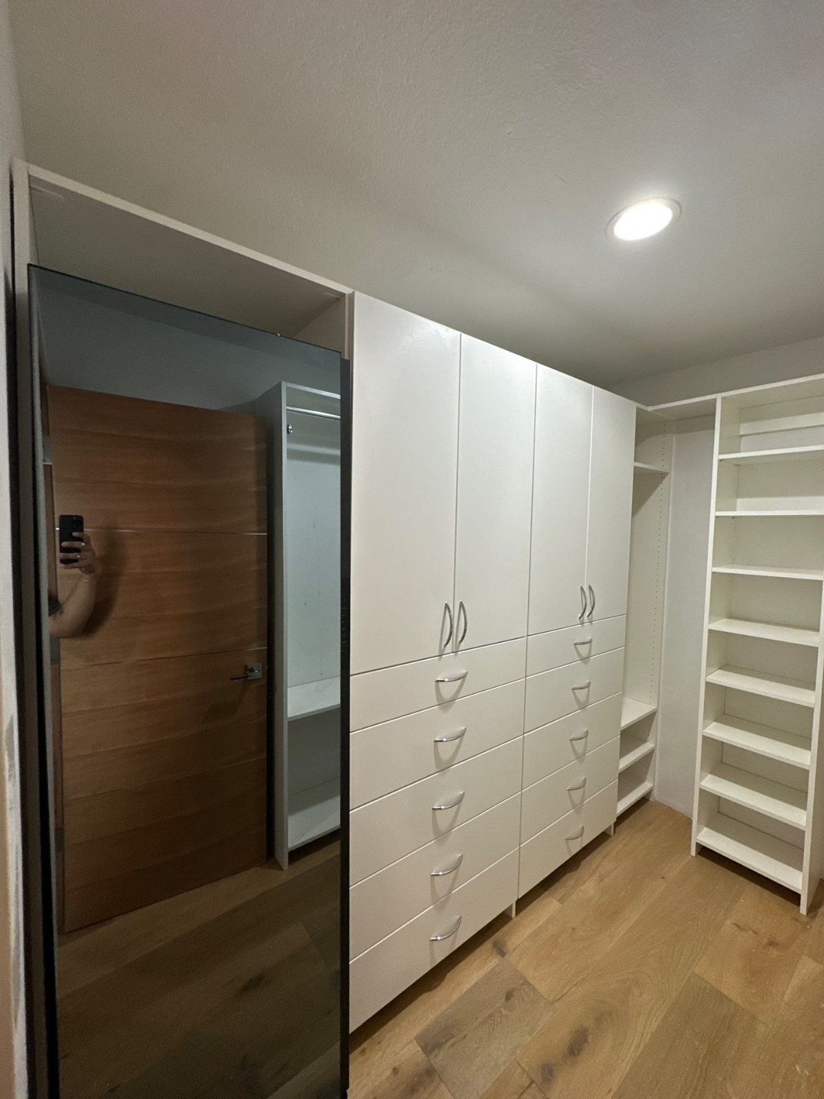 White closet with drawers and shelves; wooden floor; a large mirror; doorway.