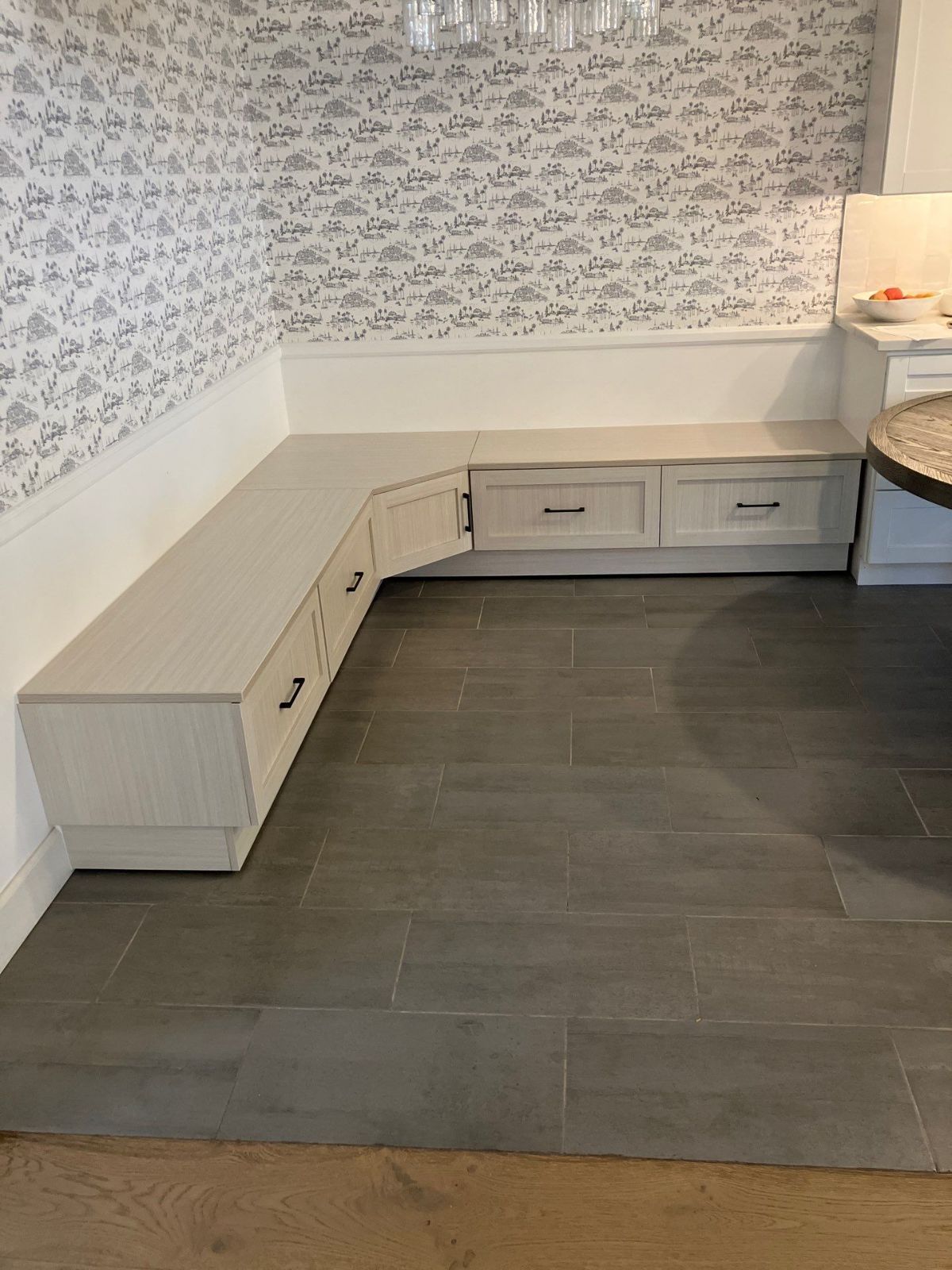 Built-in L-shaped bench with drawers in a dining area, gray floor, white and floral wallpaper, and a round table.