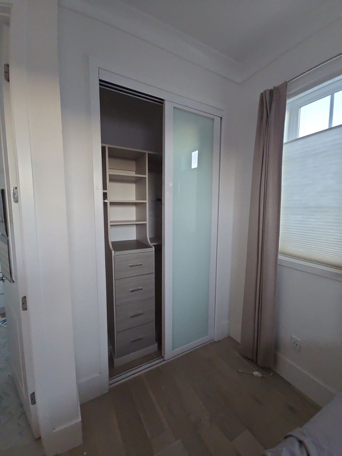 A closet with sliding frosted glass doors, a built-in shelving unit, and a window with a curtain.