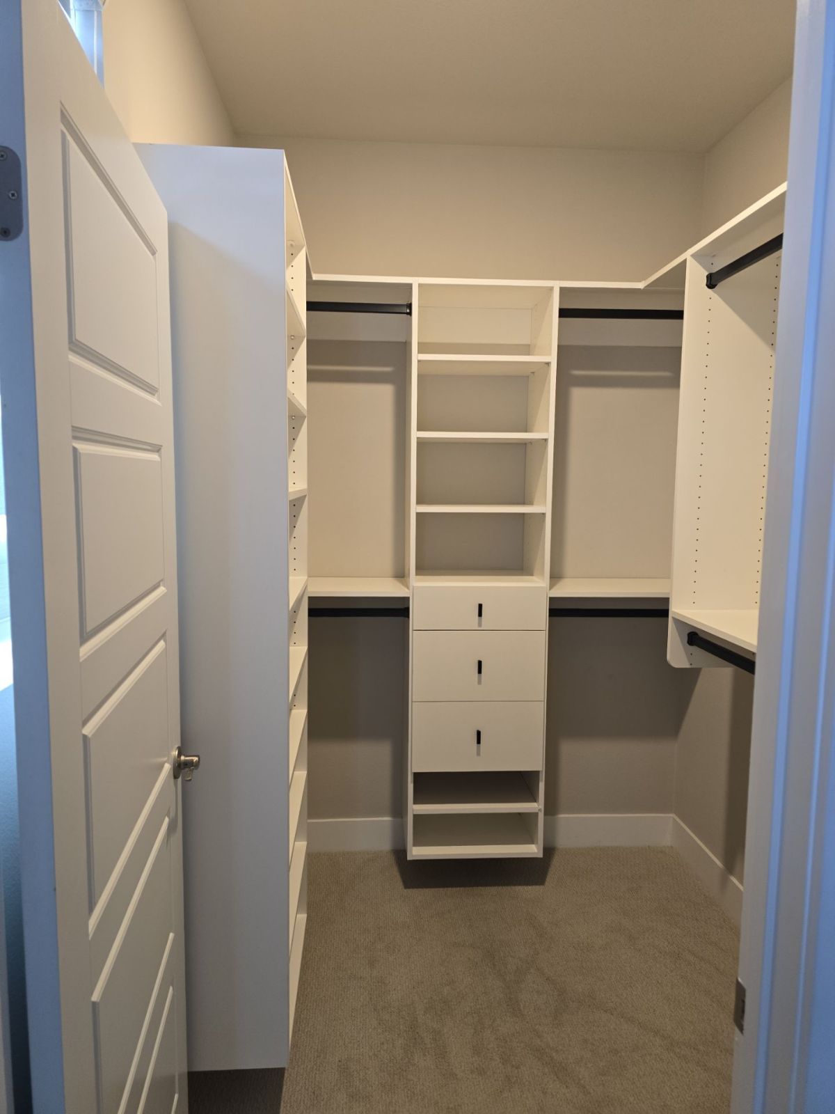 Empty white walk-in closet with shelves, drawers, and hanging rods.  Open white door on the left, grey carpet.