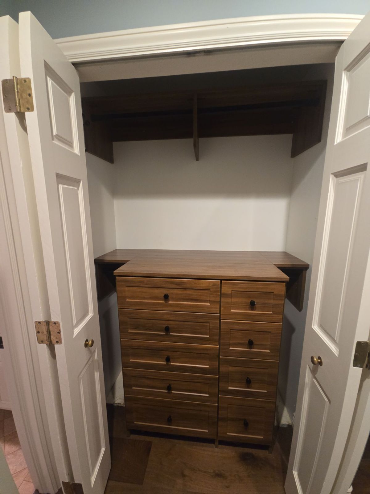 Closet with a wooden dresser, white walls, and a shelf above.