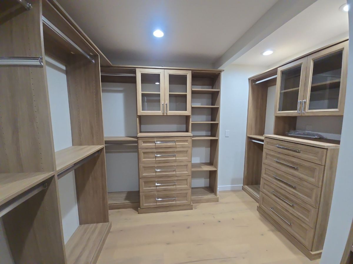 Walk-in closet with light wood shelving, drawers, and glass-door cabinets.