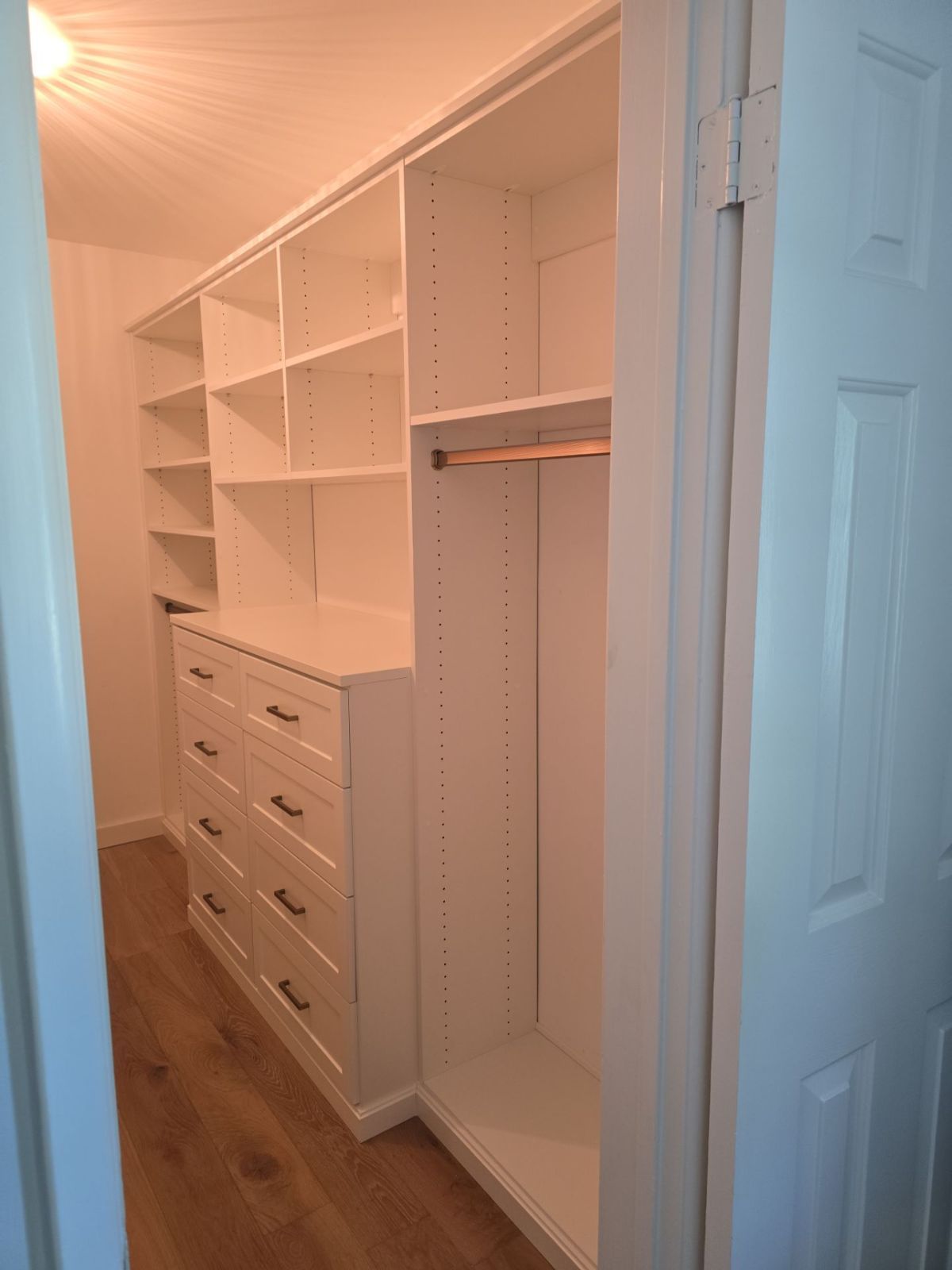 White walk-in closet with shelves, drawers, and hanging rod. Light wood-look flooring. Door on the right.