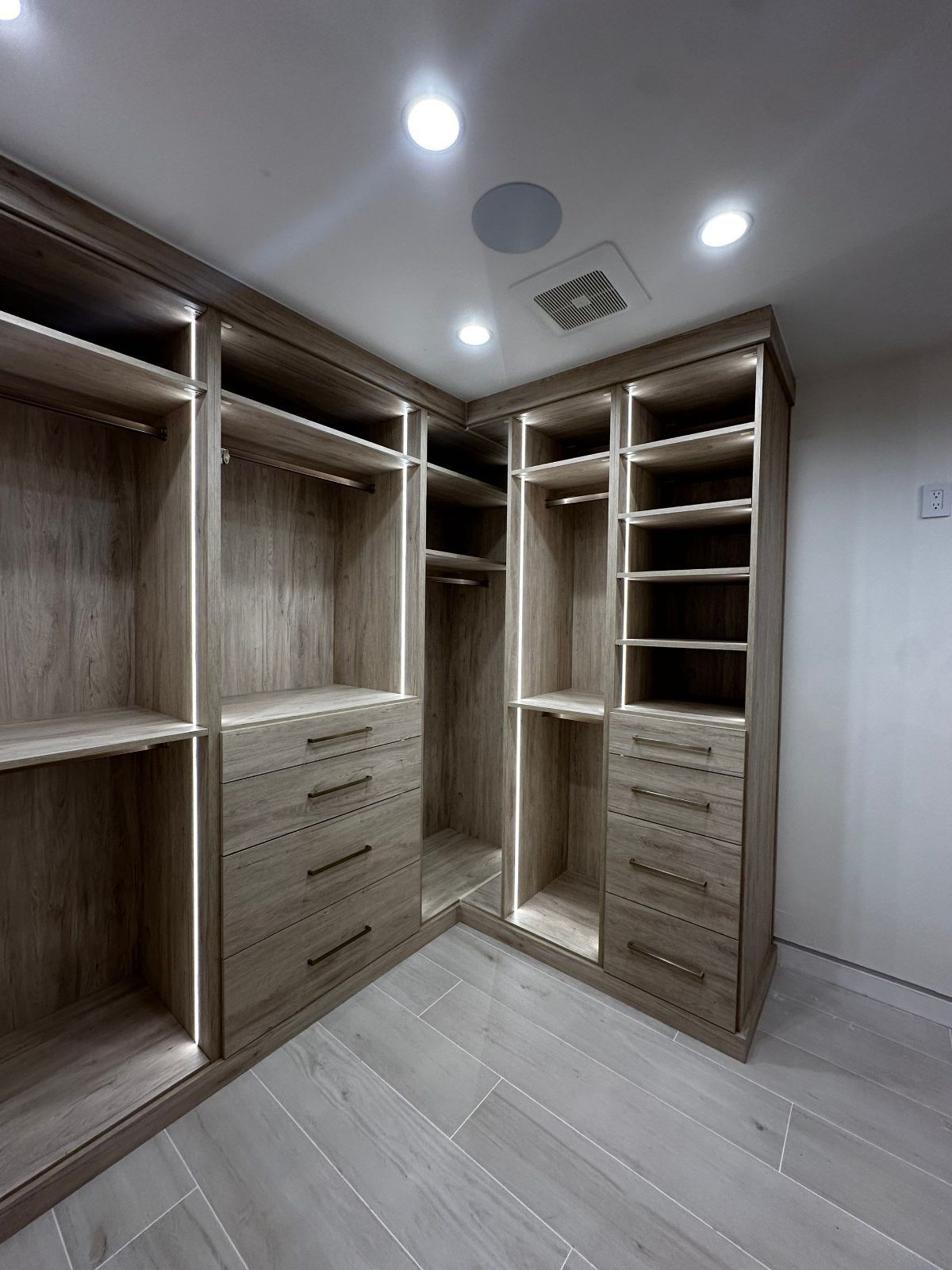 Walk-in closet with light wood shelving, drawers, and hanging rods. The corner unit has built-in lights.