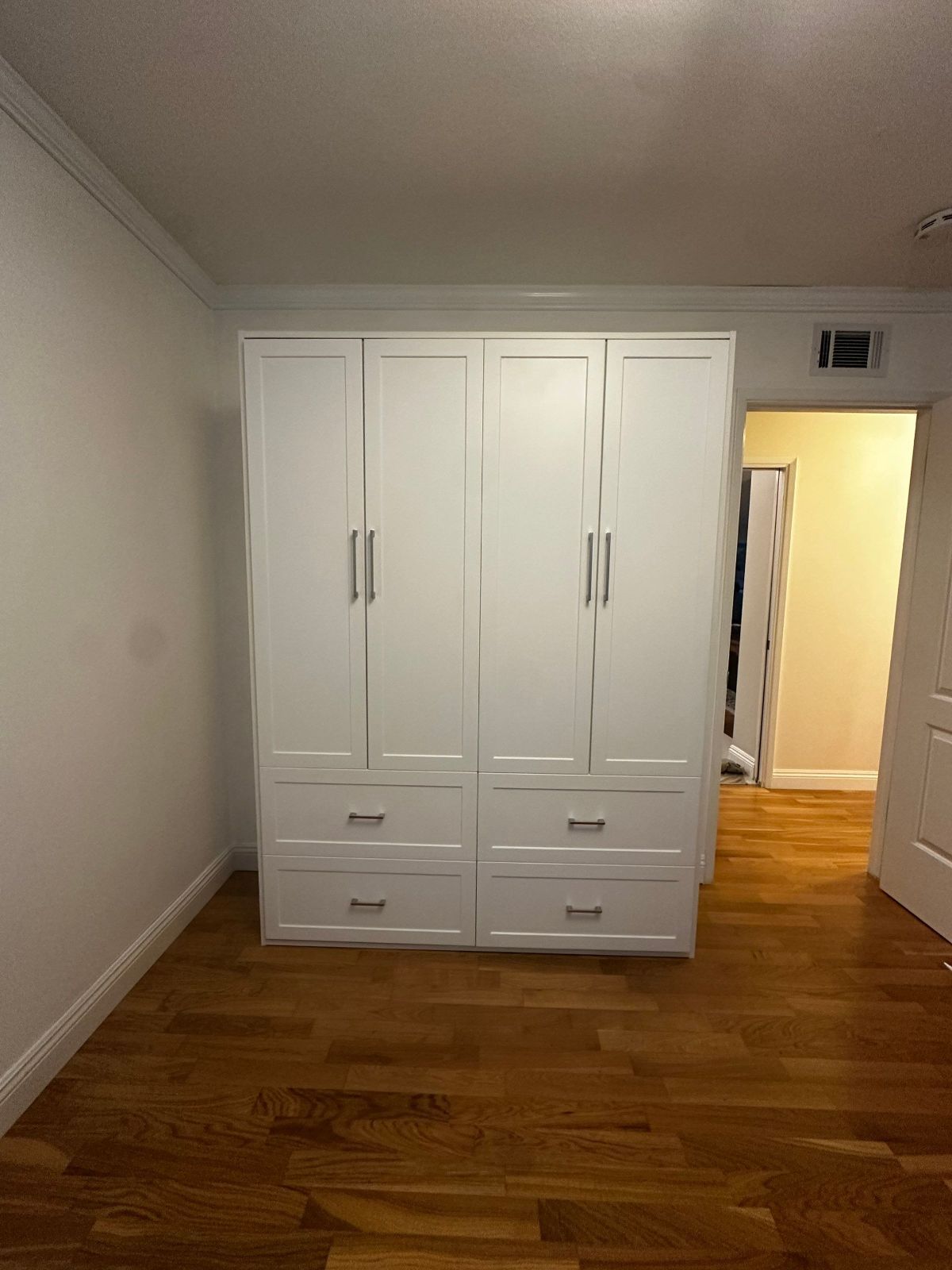 White wardrobe with four doors and two drawers in a room with hardwood floors and a doorway.