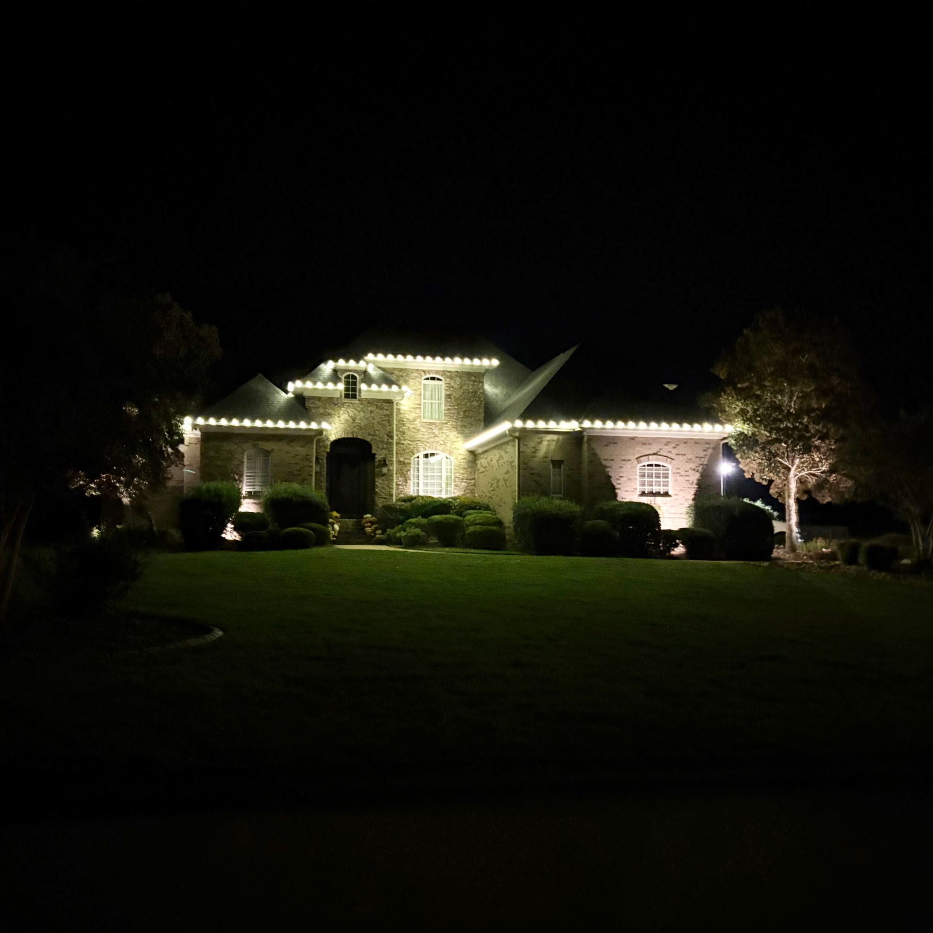 Large house at night, illuminated by white lights along the roofline and landscaping.