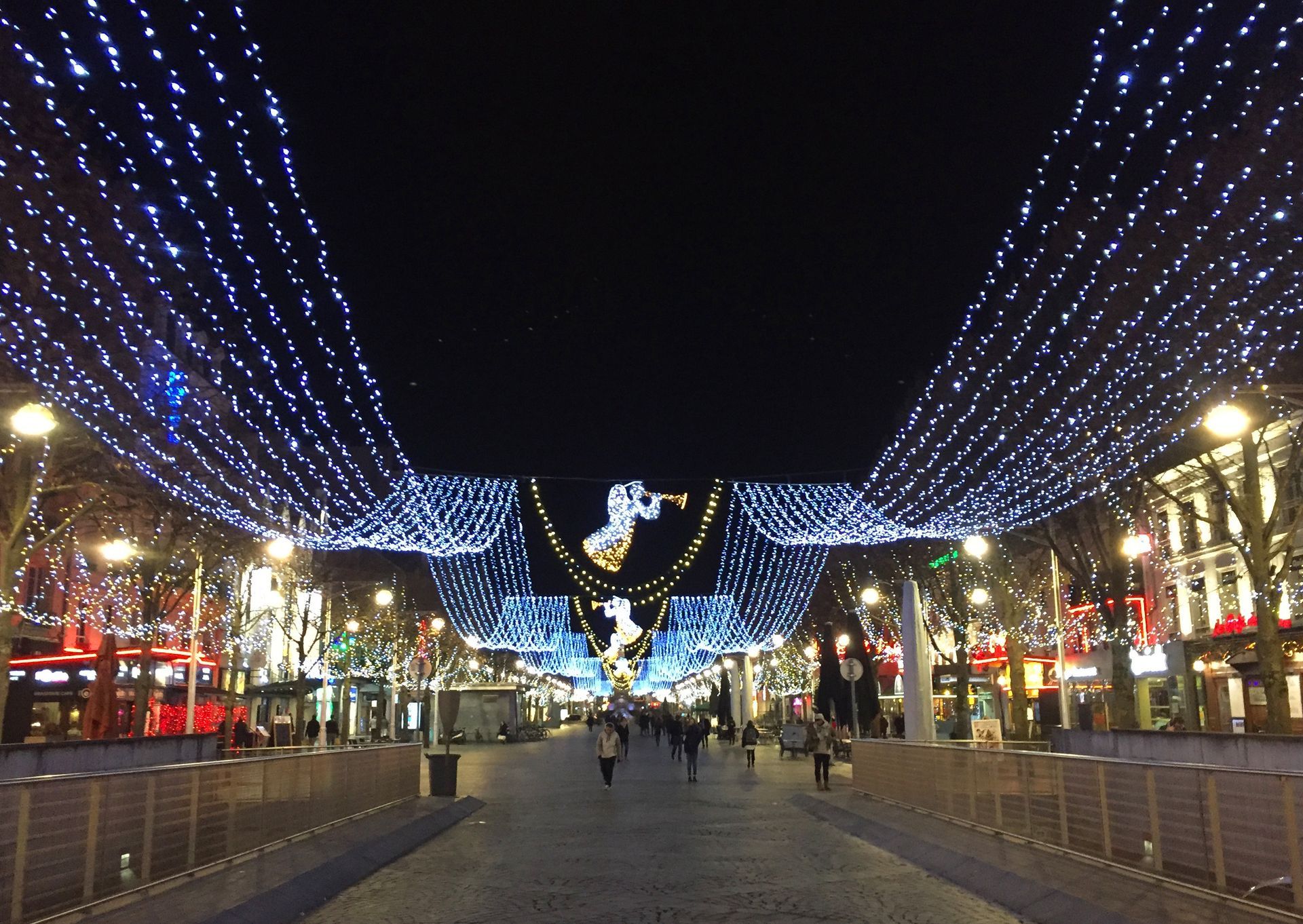 Street at night, lined with shops and festive blue Christmas lights. People walk along the street under decorations.
