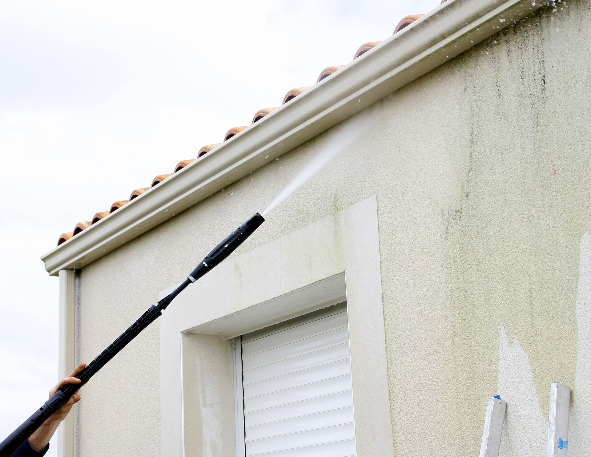 Person using a pressure washer to clean a house exterior with visible algae.