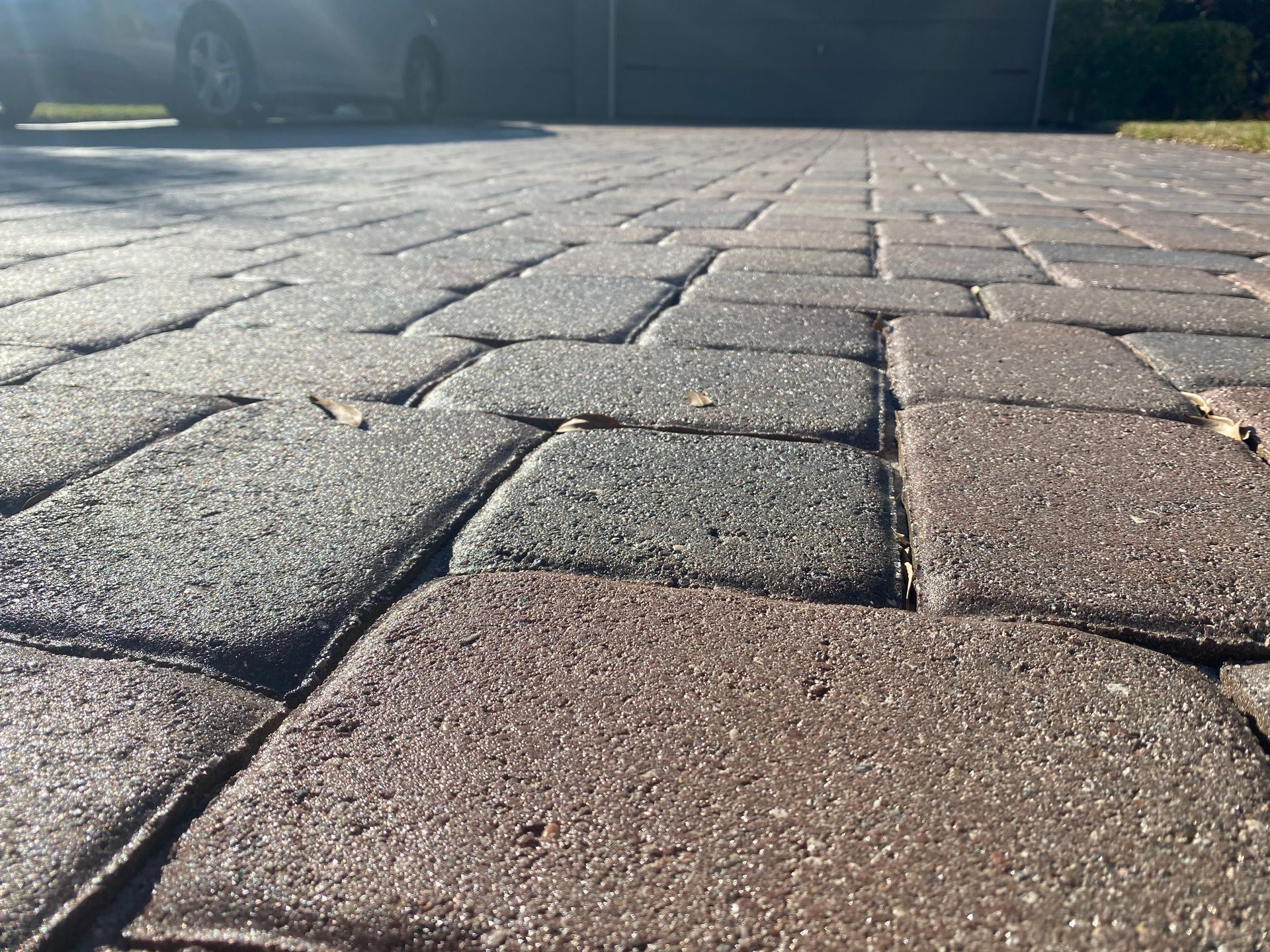 Close-up of brick pavers on a driveway, with a car in the background, lit by sunlight.