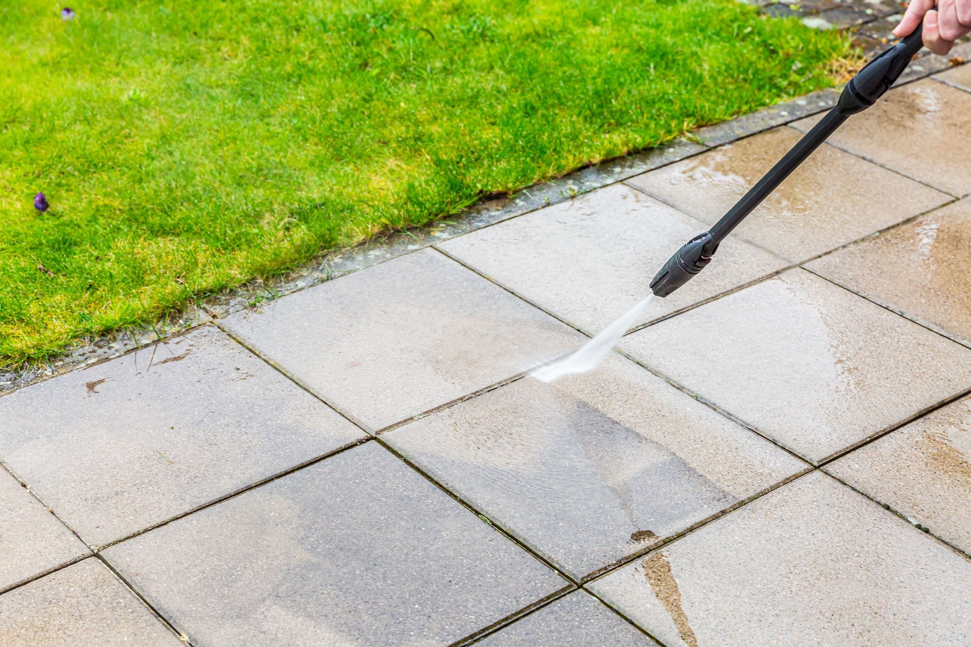 Person power washing a patio with green grass beside it.