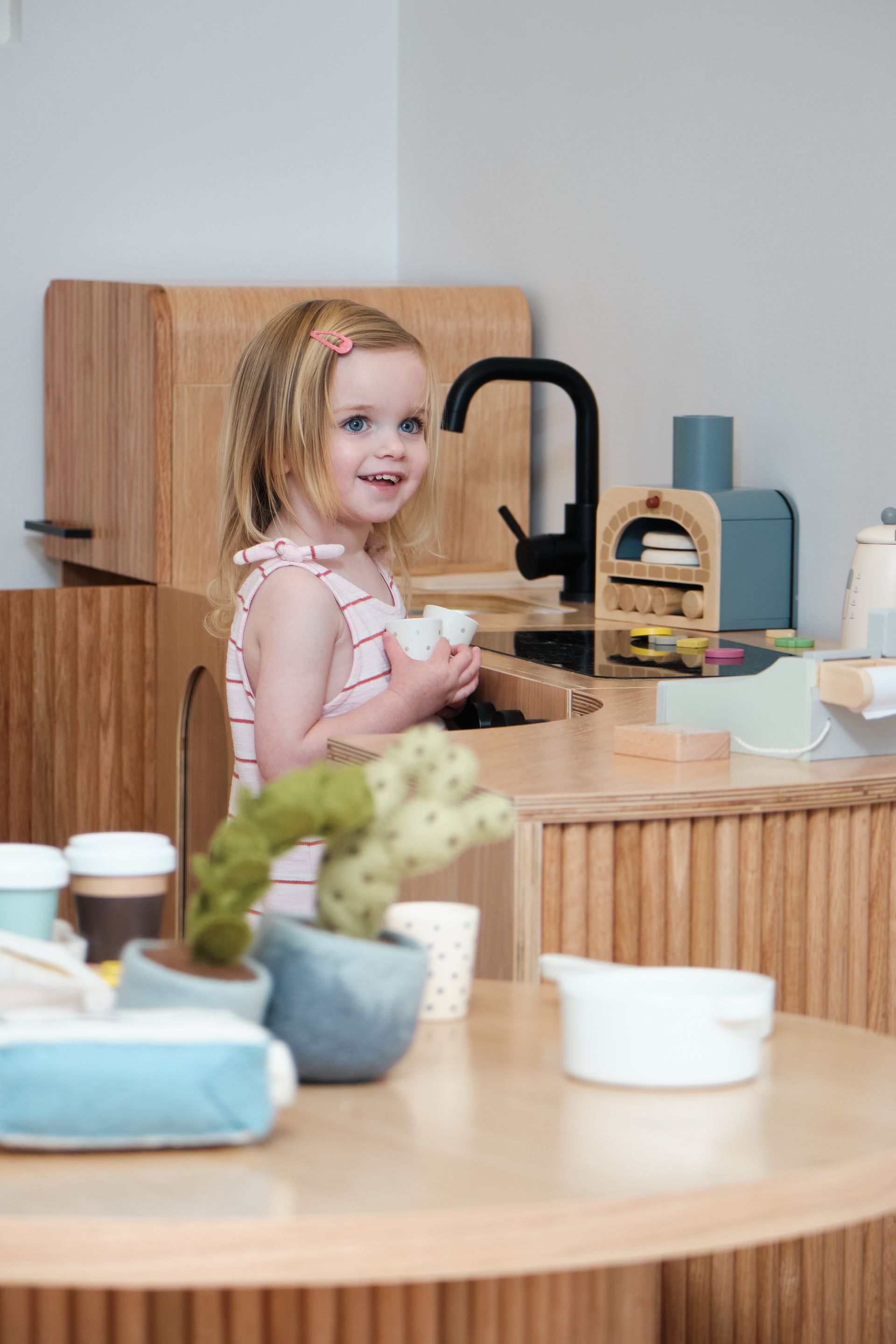 Girl playing in a wooden play kitchen, holding dishes, smiling.