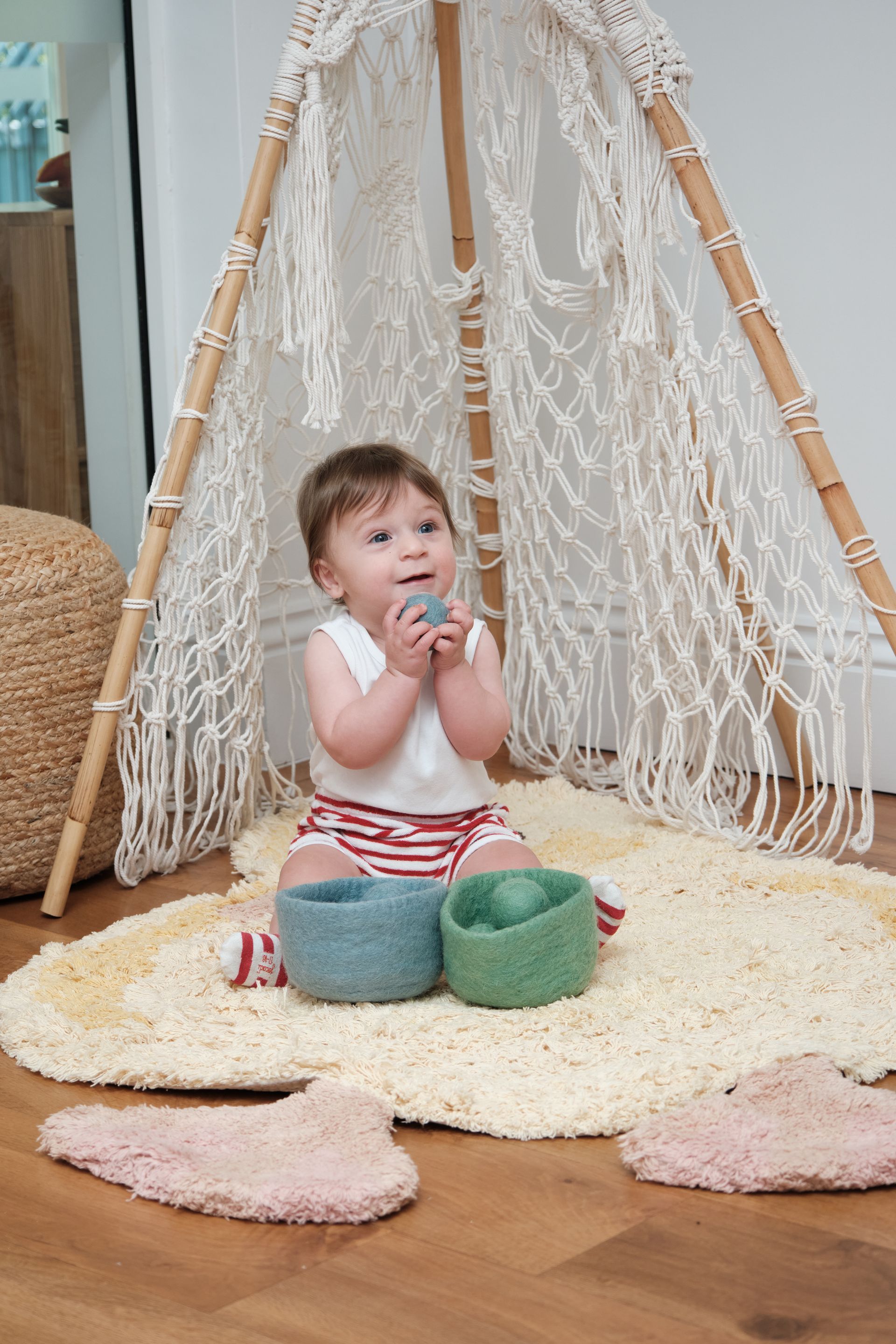 Baby playing inside a macrame teepee, holding a ball, with colorful baskets and rug.