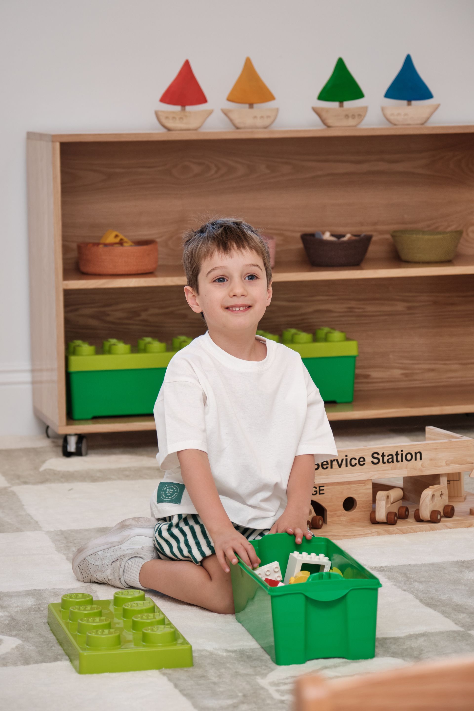 Boy sitting with green blocks, smiling, in front of a shelf with toys and boats.
