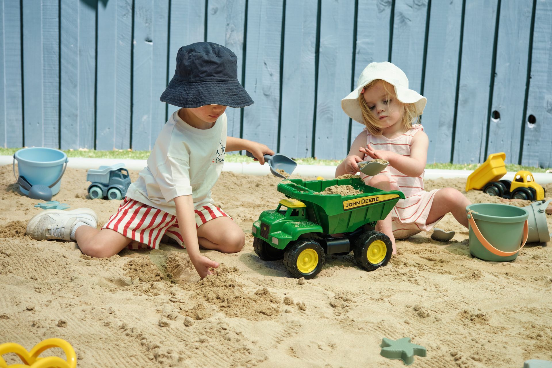 Two children in hats playing with a toy truck and sand in a sandbox.