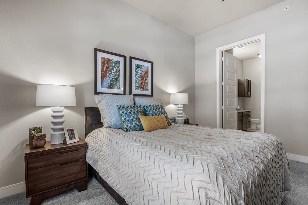 Modern bedroom interior with a large bed adorned with patterned bedding and multiple pillows, flanked by matching nightstands with white lamps. Coordinated artwork hangs above the bed, and an open door shows an adjoining bathroom in the background at Marquis at Barton Trails in Austin, TX.