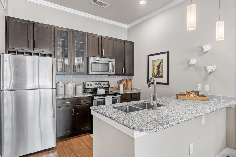 A modern kitchen with dark brown cabinets, stainless steel appliances, including a refrigerator, microwave, and stove. There's a granite countertop, light blue subway tile backsplash, and pendant lighting. Decor includes wall-mounted bird sculptures and a framed picture. A kitchen island features a sink and a cutting board with a baguette at Marquis at Barton Trails in Austin, TX.