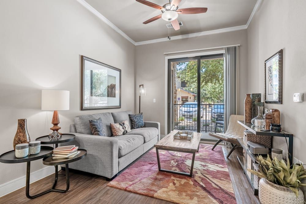 A cozy, well-decorated living room interior with a neutral color palette. It features a comfortable grey sofa adorned with patterned cushions, a wooden coffee table, and a side table with decorative items and books in the foreground. A floor lamp and table lamp provide soft lighting. A colorful area rug adds a touch of warmth to the hardwood floor. The room is completed with framed artwork on the walls and a sliding glass door in the background that opens to a balcony with an outdoor view, allowing natural light to fill the space at Marquis at Barton Trails in Austin, TX.