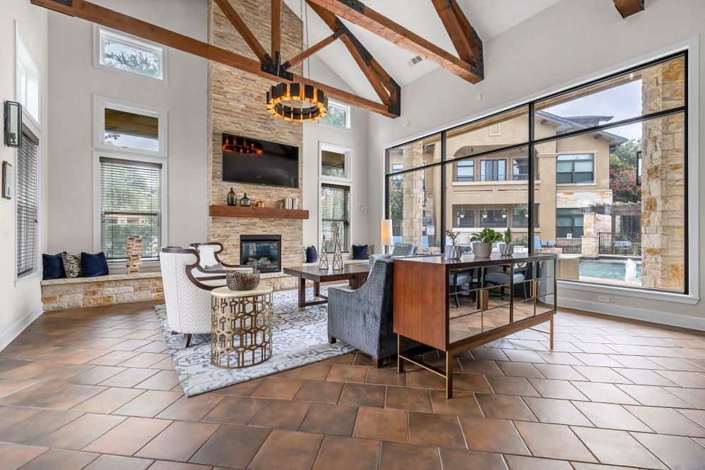 Spacious modern clubhouse with high ceilings, exposed wooden beams, and a stone fireplace. Elegant seating area with a patterned rug, armchairs, and a sofa faces a mounted TV. Large glass windows provide abundant natural light and a view of the outdoor pool area. An adjoining dining space features a wooden table set and stylish chandelier. The room is finished with terra cotta tiled flooring at Marquis at Barton Trails in Austin, TX.