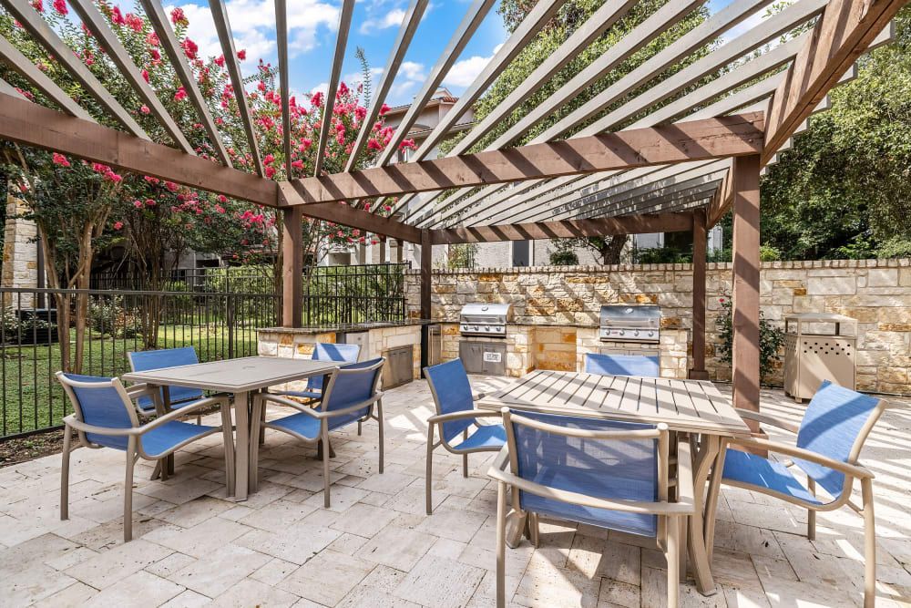 An inviting outdoor patio area with a wooden pergola draped with flowering vines, two tables with blue chairs, and a built-in cooking station with a grill and counter space. The area is enclosed by a stone wall and metal fence with greenery in the background on a sunny day at Marquis at Barton Trails in Austin, TX.