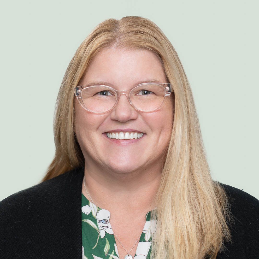 A woman wearing glasses and a green floral shirt is smiling in front of a painting.