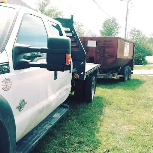 White truck with a flatbed trailer towing a rusty dumpster on a grassy area.