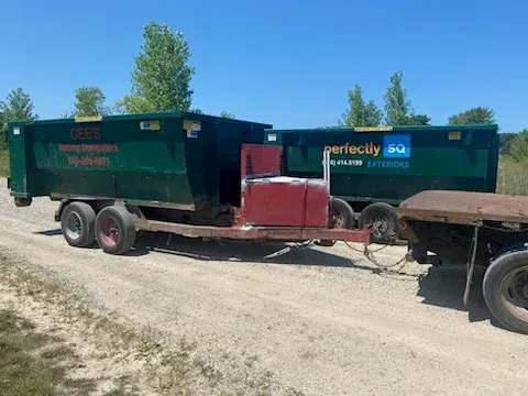 Two dark green dumpster trailers hitched together on a gravel road, under a blue sky.
