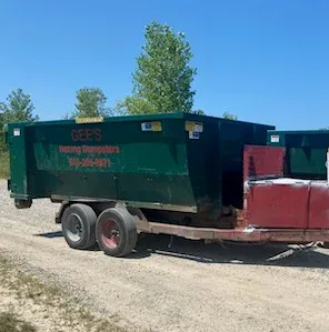 Two dark green dumpster trailers hitched together on a gravel road, under a blue sky.
