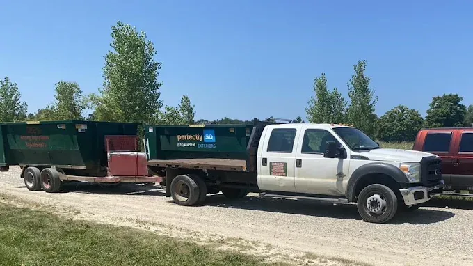 White flatbed truck pulling a trailer with a green container on a gravel road.