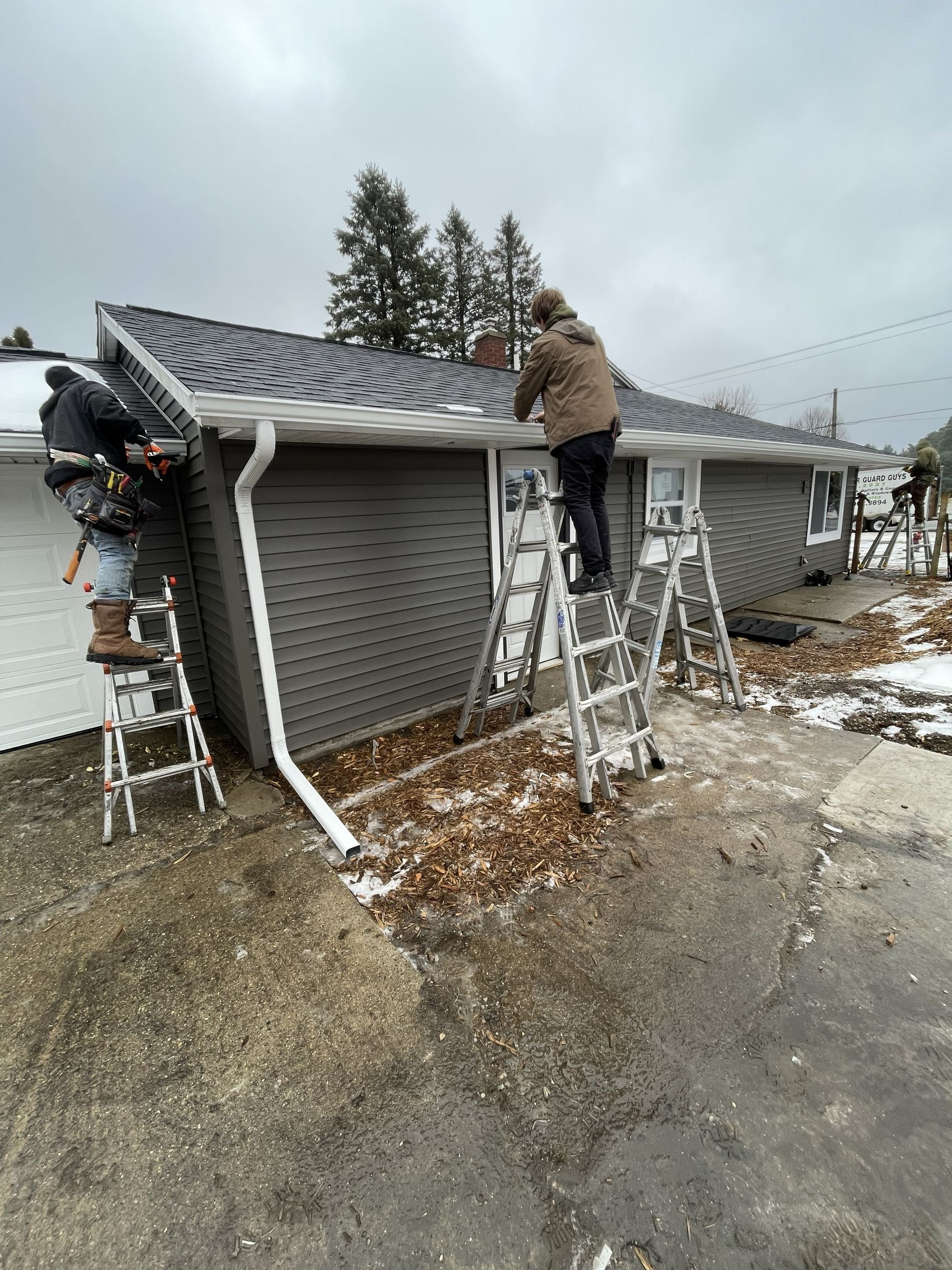 Beige house siding with white trim, gutter, and fall foliage in background.