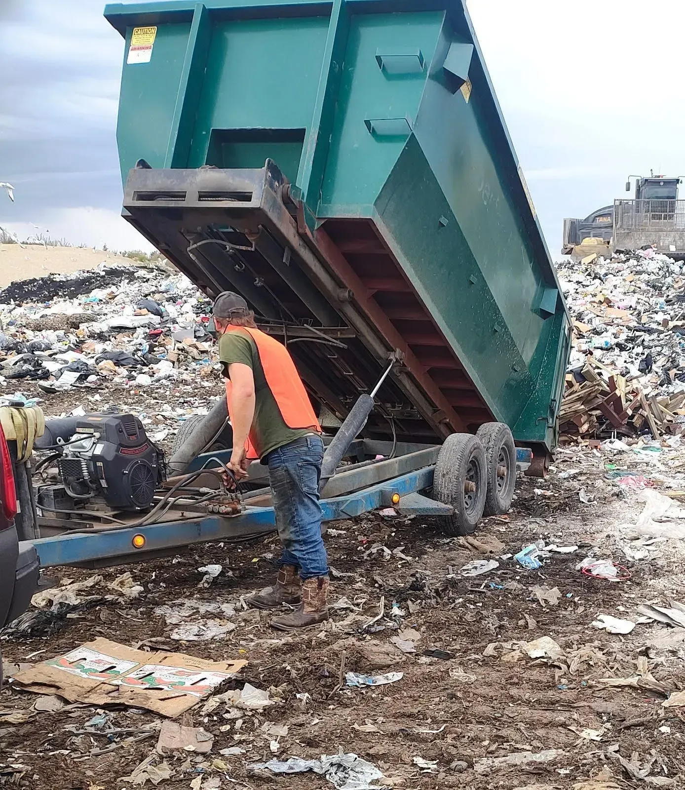 A person in an orange vest, working near a raised, green trailer dumping waste at a landfill.