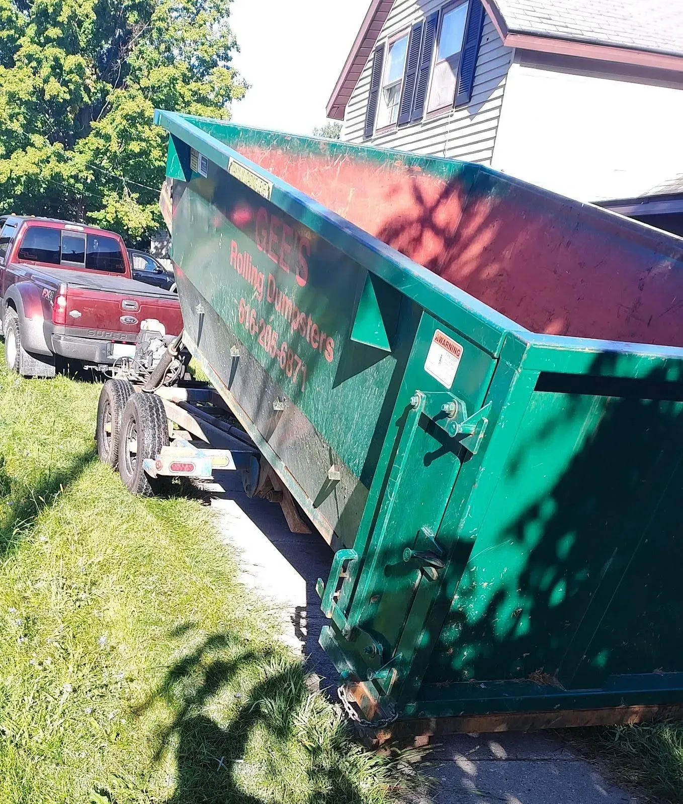 Green dumpster on a trailer, parked on grass next to a sidewalk and a red truck.