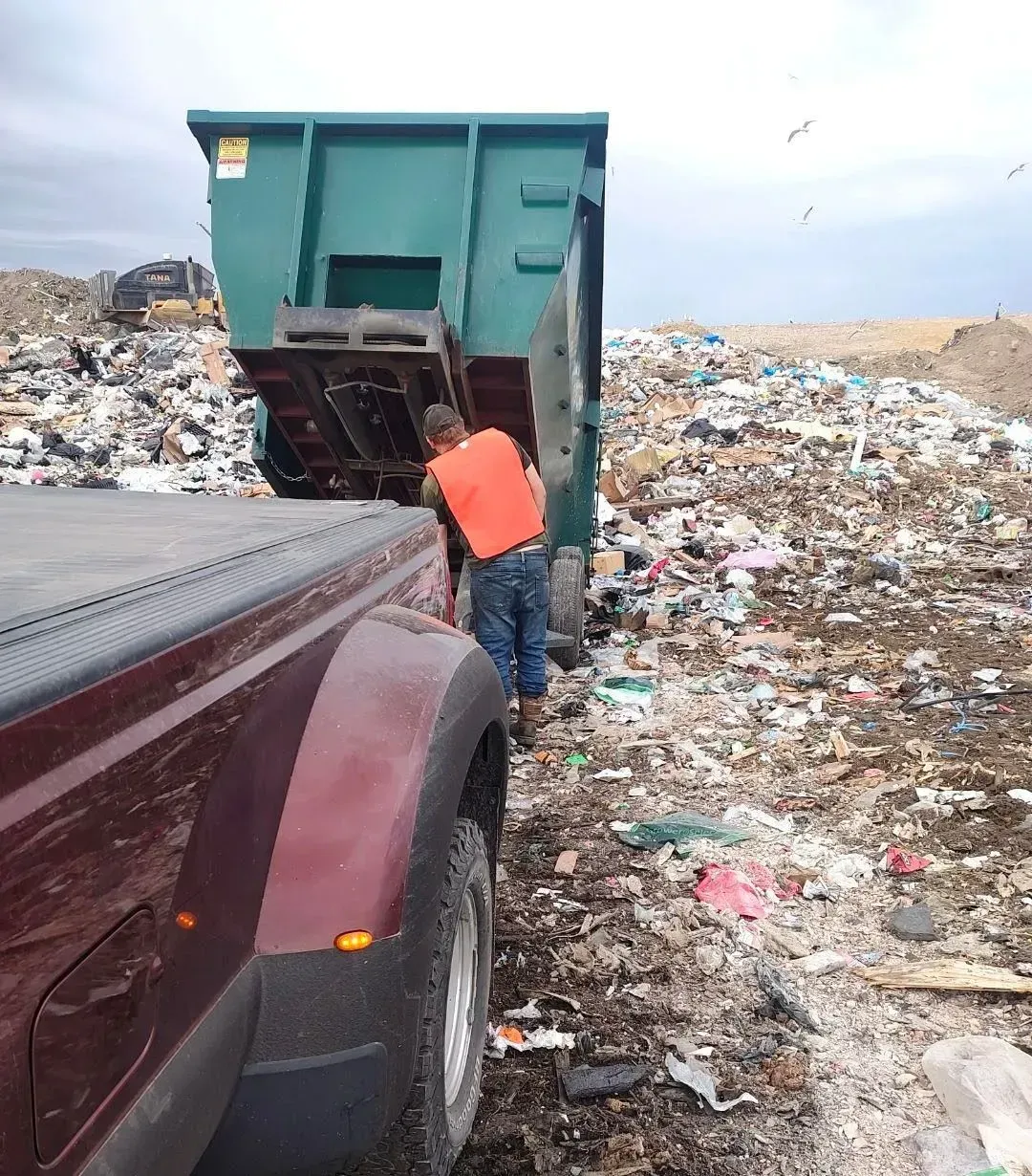 A worker in an orange shirt inspects a dumpster being emptied at a landfill. A maroon truck is in view.