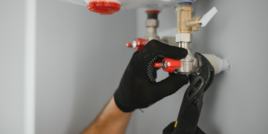 Plumber in black gloves uses a wrench on water pipes against a grey wall.