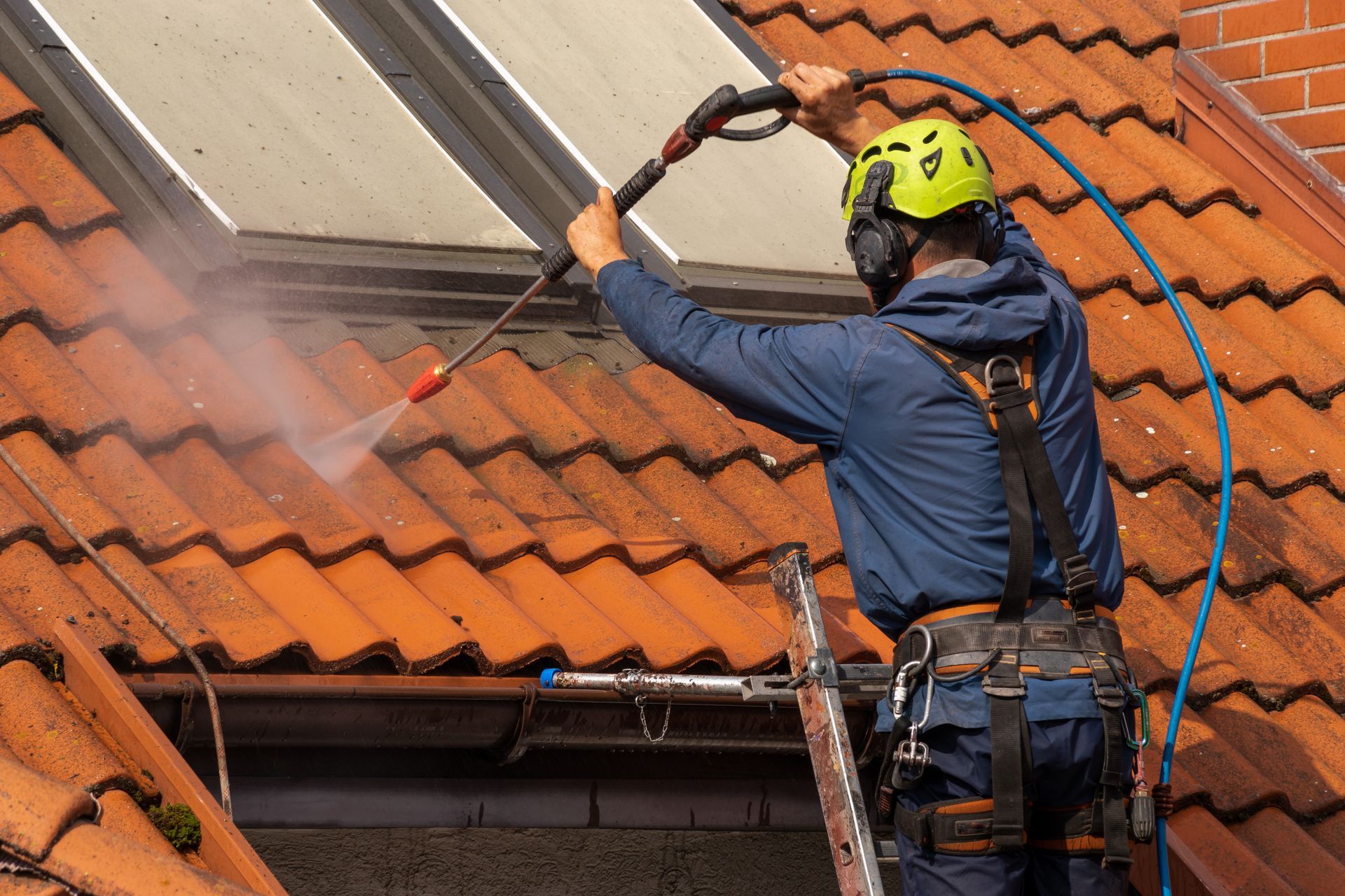 Person on a ladder power washing a tiled roof, wearing a safety harness and helmet.