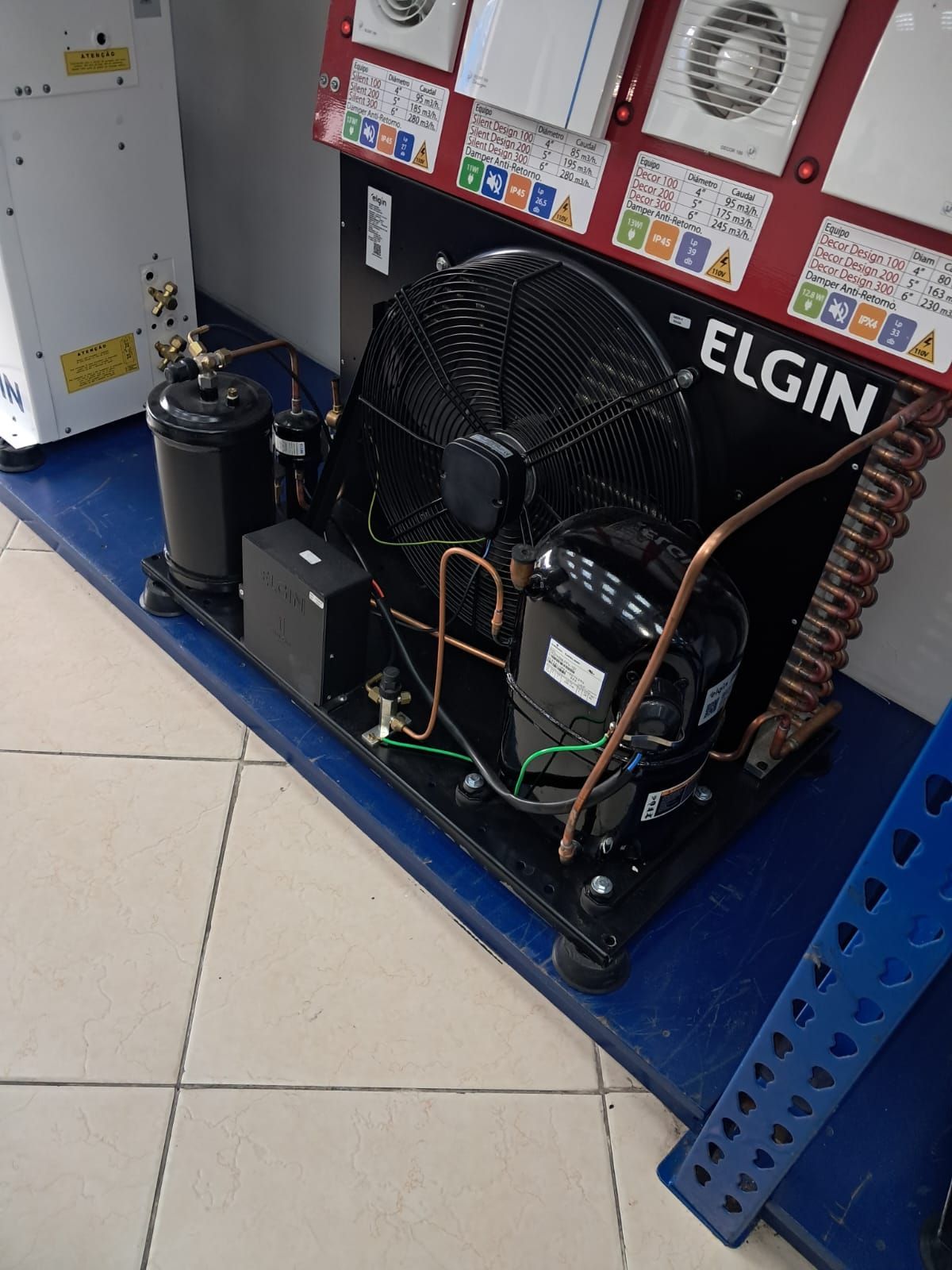 Unidad de refrigeración negra con ventilador, tubos de cobre y componentes sobre una superficie azul.