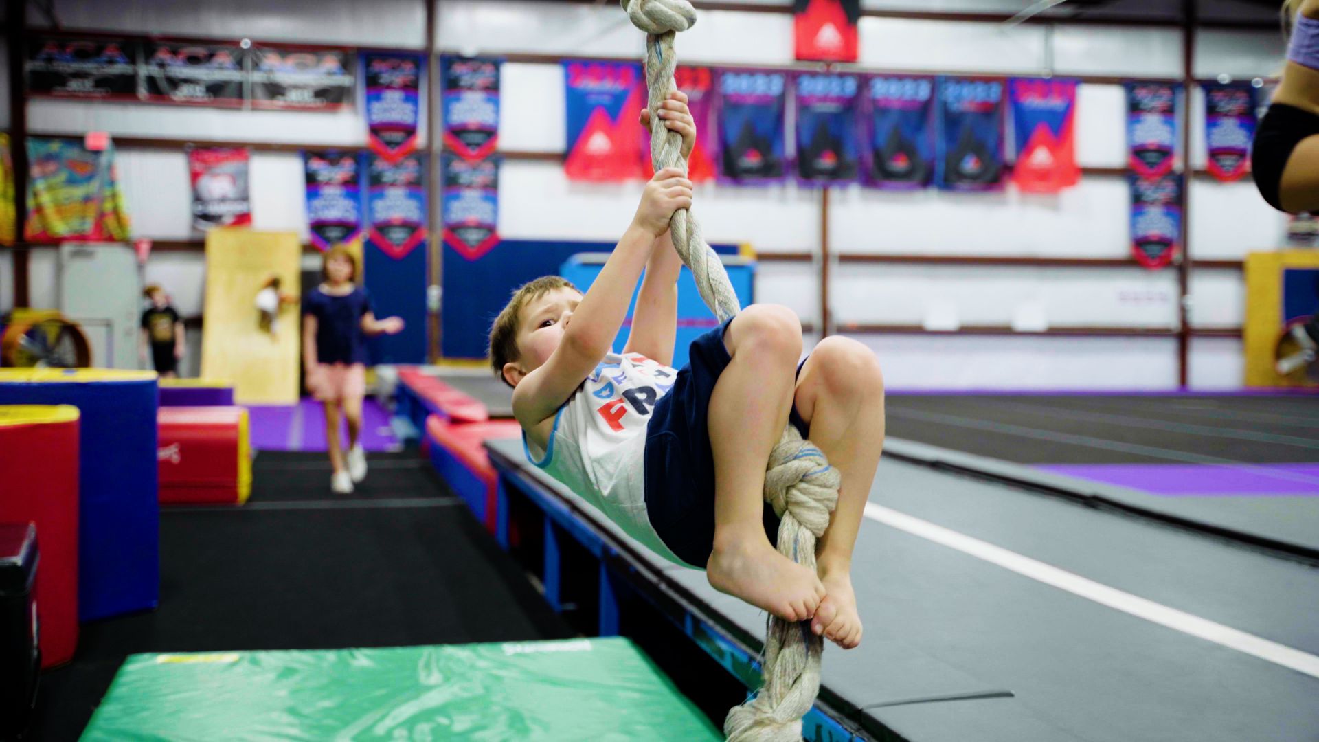 A young boy is climbing a rope on a balance beam in a gym.