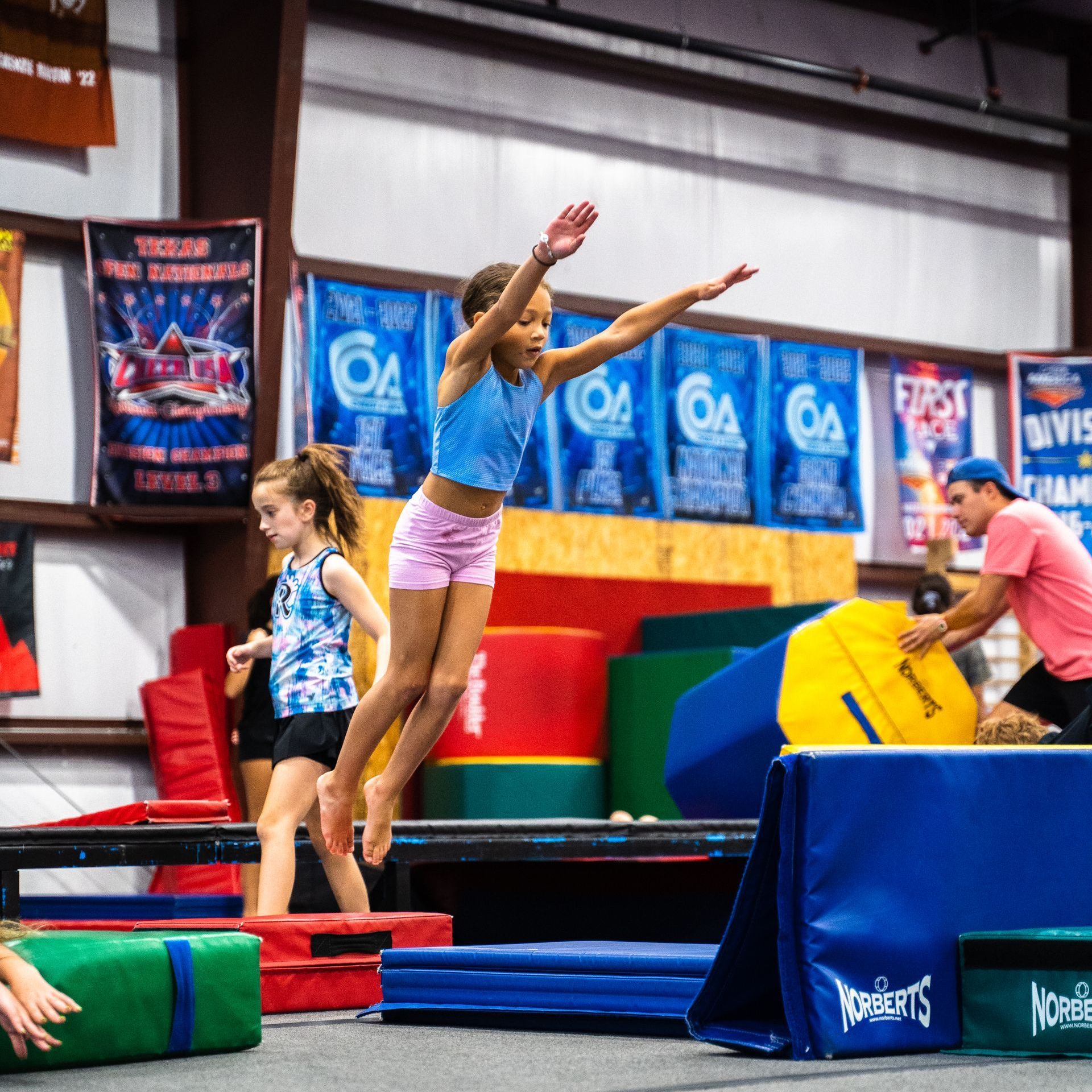 A girl in a blue top and pink shorts is jumping in the air