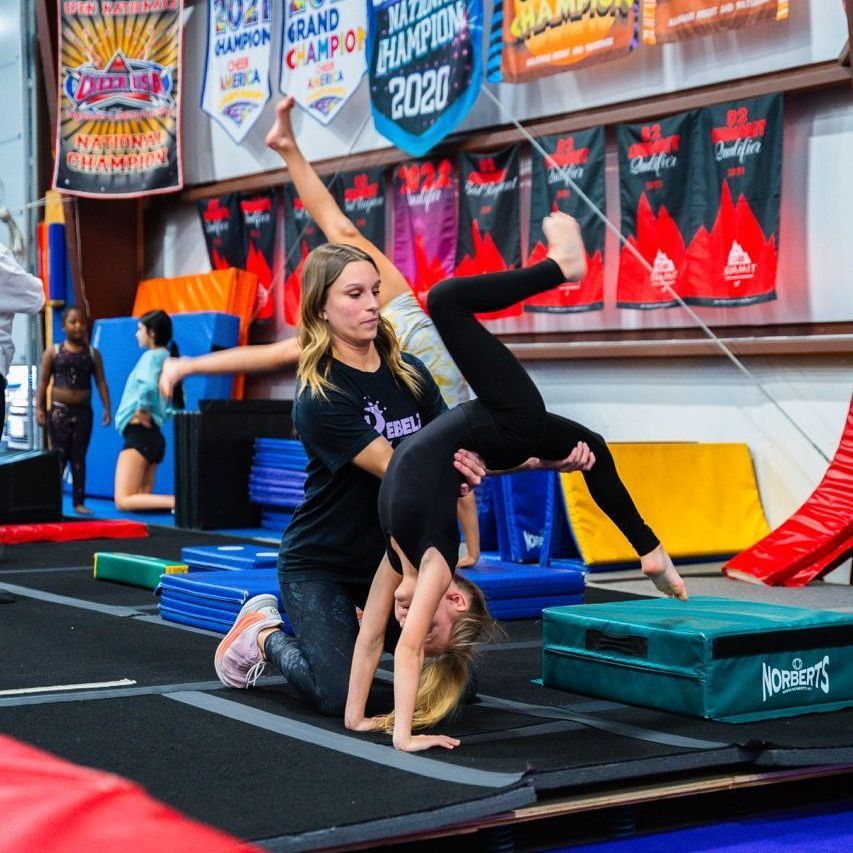 A woman is helping a young girl do a handstand in a gym